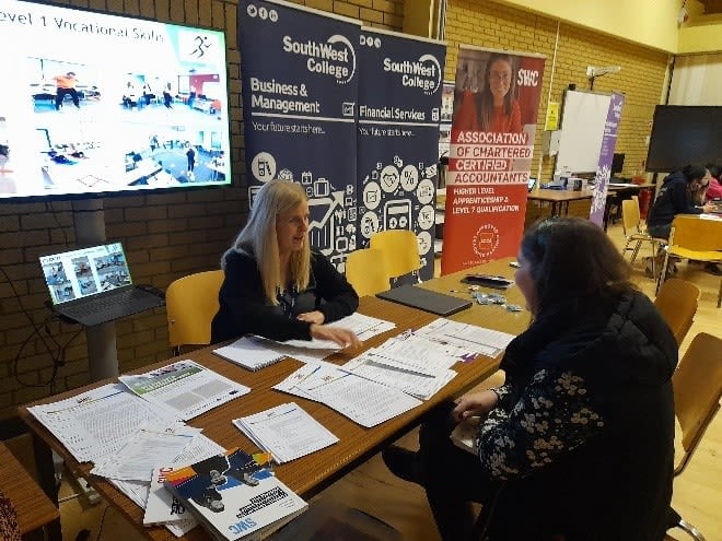 Image shows two people sitting opposite each other at info desk. Leaflets cover the desk and one person is leaning forward explaining to the other.