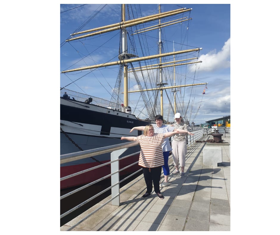 Three service users standing beside a sail ship with arms raised.