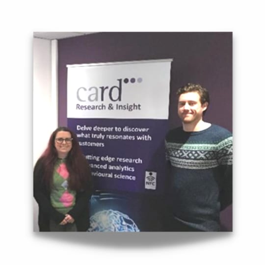 Images shows two people standing either side of a banner that reads card. One taller than the other. Both brown hair.